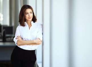 Женщина в деловом костюме уверенно смотрит в даль, скрестив руки на груди.